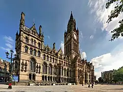 A grand gothic revival building as viewed from afar, with a gothic revival clock tower stretching high into the sky. Trees partially obstruct the view and the sky is clear with some clouds.