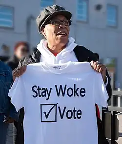 Congresswoman Marcia fudge at an outdoor gathering holding a T-shirt in front of her which reads "Stay Woke" followed by a tick mark inside a box and the word "Vote"
