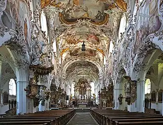 A symmetrical view straight down the aisle of a highly ornate church interior with towering white columns, extensive pastel and gold stucco decorations, and a vaulted ceiling covered in elaborate frescoes.