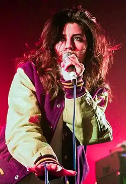A young brunette woman wearing a baseball-style jacket, singing into a microphone against a pinkish background