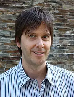 alt=Portrait photo of a white man with dark brown hair wearing a striped dress shirt.