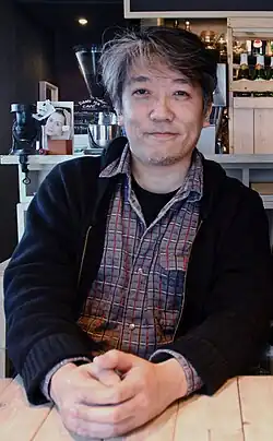 Portrait of a Japanese man sitting at a table wearing a black jacket and grey flannel shirt.