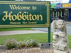 A freestanding green sign reading "Welcome to Hobbiton" in large gold calligraphic letters, with "Matamata New Zealand" appearing under it in smaller letters. In front of the sign is a statue of a grotesque on a pedestal. A lawn, road, and shops are visible in the background.