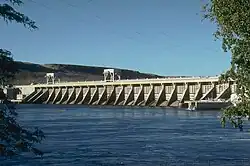 A concrete hydroelectric dam's spillway on the downriver side. Water is in the foreground and power lines run overhead.