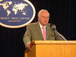 A white-haired man in a suit and pink tie is standing at a lectern, speaking into a microphone. Behind him is a plaque reading "FOREIGN PRESS CENTER WASHINGTON".