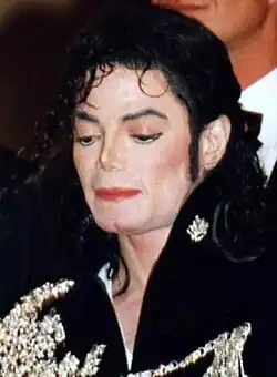 Jackson is wearing sun glasses and a black jacket with a golden metal rectangle on both sides of the collar, golden badges on both upper arms and a golden armband on his right arm under the badge. Two golden bandoliers cross his torso forming the letter "X". A third bandolier runs from his left shoulder to the bucket of his golden belt. He is frozen. His head is turned to the left. His hair is falling in his eyes and he is wearing a headset microphone. His skin is light