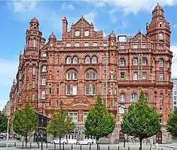 A large red-brick building with curved windows as viewed from a short distance away. There are green trees in front and a blue sky behind. The building curves round on both sides to follow the road.