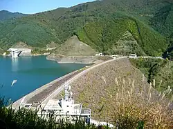 A large rocky dam, with a reservoir behind it, and green hills in the background.