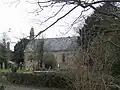 Stone building partially obscured by trees. In the foreground are gravestones.