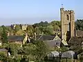 View of the roofs of houses with a prominent square church tower, interspersed with trees.