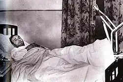 A man lies in a bed with his left leg raised by a pulley.