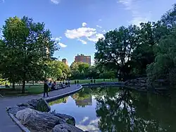 A pond in the foreground with a path on its shore and high-rise buildings in the background