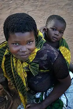 A young mother smiles up at the camera. On her back is her baby, gazing at the camera with lively interest.