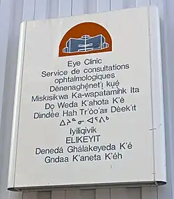 A metallic white sign on a grey background with a red and blue depiction of a building at the top. Below it is text in blue saying "Eye Clinic" in English, French and the other nine official territorial languages