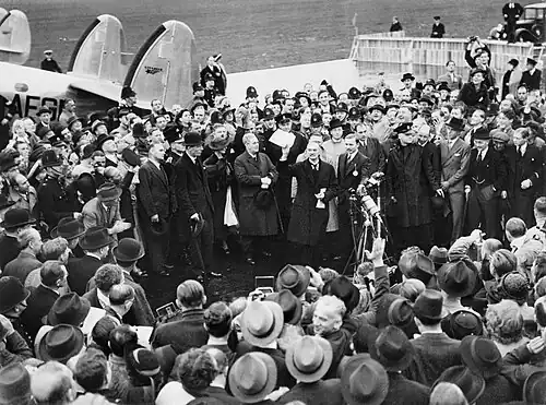 A large crowd on an airfield; British prime minister Neville Chamberlain presents an assurance from German chancellor Adolf Hitler.