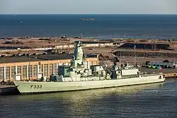 Photograph of NRP Bartolomeu Dias (F333) of the Portuguese Navy docked by the quays of Hernesaari, Helsinki, Finland, with the Portuguese flag flying at the stern and calm blue water stretching out to the open sea beyond.