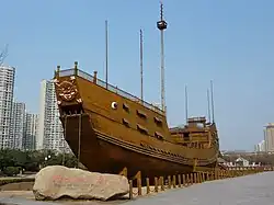 Large wooden replica of a traditional Chinese treasure ship displayed on land, with a carved dragon prow, surrounded by a walkway and modern high-rise buildings in the background under a clear sky