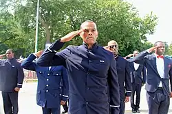 A group of black men, with closely cropped hair and wearing navy blue suits, in a line saluting