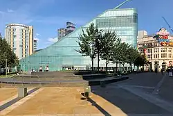 A triangular prism shaped glass building as photographed from a short distance away, with trees lined in front.