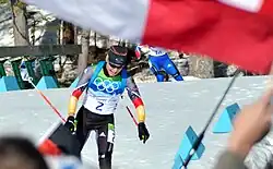 A woman in multicoloured winter sportswear which features the Olympic rings on her chest and the number 2 in the center, moves towards the camera. She is pictured in an area covered in snow. A second person can be seen in the background to her right. An out of focus red-and-white flag covers the upper parts of the image.