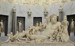 A large marble sculpture, inside a formal room. The main object is a reclining nude man, surrounded by many children, as well as some animals.