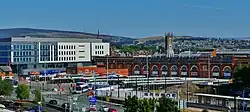 A panoramic shot of a town. In the foreground a modern-looking bus station in front of a red-brick building with large sash windows; in the background a church spire and a hill with houses on the hillside.
