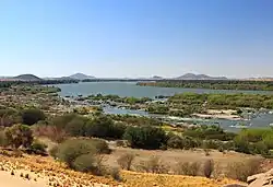 A wide, calm river, under a blue sky, with many bushes on its sandy banks.
