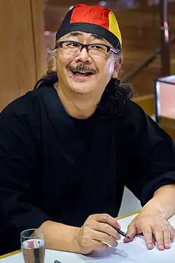 Portrait of a mustached Japanese man wearing glasses and a black, red, and yellow head bandana.