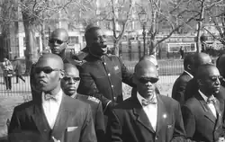 A group of young black men, wearing suits and with shaven heads, standing outdoors. One is in the center, speaking aloud. The others surround him, facing outward. Although the speaker does not wear sunglasses, the others all do.