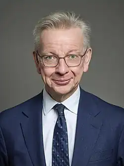 Gove facing forward in a blue suit jacket, white shirt and blue tie against a grey background