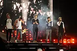 Five young adult males stand next to each other on a stage, all wearing jeans and long-sleeve t-shirts, except for band member Liam Payne, who wears a tank top. From left to right: Louis Tomlinson, Liam Payne, Harry Styles, Zayn Malik, and Niall Horan.