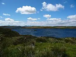 The wind creates ripples on a blue lake surrounded by a low-lying green and brown landscape under small white clouds in a blue sky. Mountains line the distant horizon.