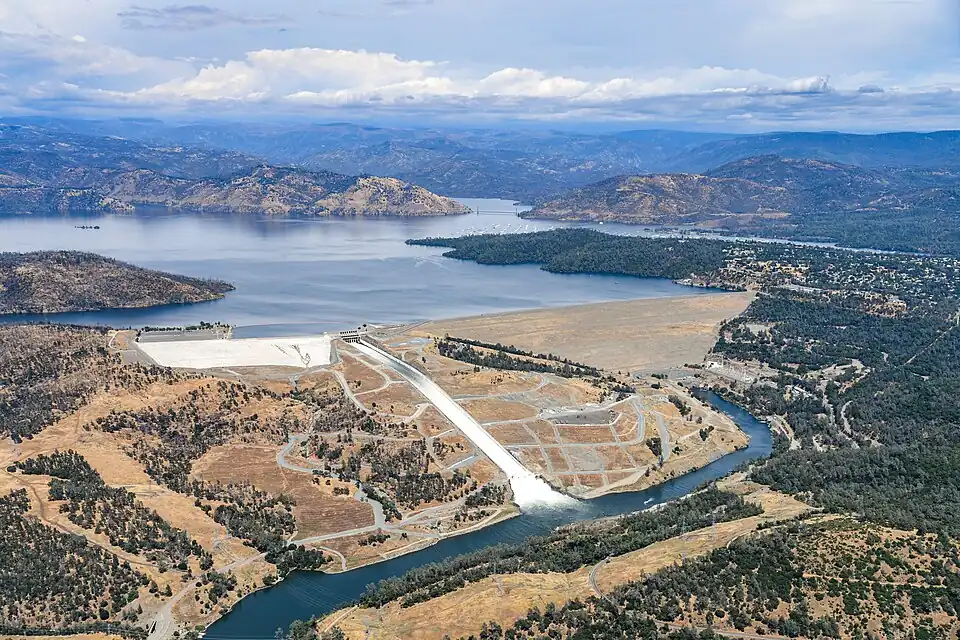 A broad, gently sloping dam made of rocks impounds a lake. Water is spilling downhill next to the dam, within a concrete channel.