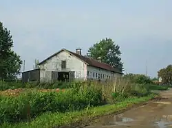 View of a long red-roofed farm building surrounded by overgrown ground at the side of a muddy road
