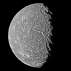 A round spherical body with its left half illuminated. The surface has a mottled appearance with bright patches among relatively dark terrain. The terminator is slightly to the right from the center and runs from the top to bottom. A large crater with a central pit can be seen at the terminator in the upper half of the image. Another bright crater can be seen at the bottom intersected by a canyon. The second large canyon runs from the darkness at the lower-right side to visible center of the body.