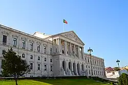Photograph of São Bento Palace. A large white neoclassical government building with tall columns and sculpted pediments rises above a green lawn under a clear blue sky, with the Portuguese flag flying from the roof.