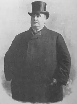 A large man stands wearing a full-length coat and top hat.