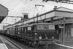 A black and white photo of a locomotive at a two-platform station. The locomotive, and especially its pantograph, look much more continental than British