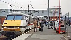 A colour photo taken from an island platform, but only one side is really visible due to people and a platform sign. On the left side is ani Intercity 225, with the photographer at the Class 91 end, and it is painted in Swallow livery. Most the length of the train is concealed by people and the station building/overbridge.
