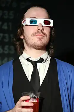 Fish wears his medium-long hair off his face, some stubble facial hair, 3D glasses, a white dress shirt with loosened black tie under a black vest and a blue cardigan while holding a red drink at a party.