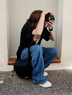 A person holding and looking through a camera sits on the stone steps of a door, pointing the camera towards the upper right