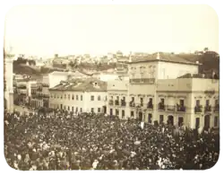 An old photograph showing a crowded square in front of a large, white, multi-storied building