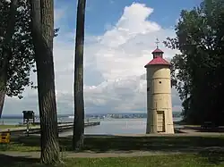 A small off-white lighthouse with a red roof and the trunks of two trees overlooking a bay with a city in the background.