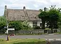 Thatched stone house surrounded by trees. In the foreground a road junction and sign.
