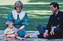 Diana in a green polka dot dress, Charles in a suit, and a baby William sit on a patterned rug outdoors.