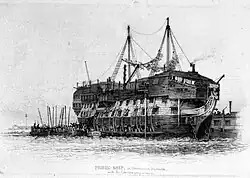 Drawing of the hull of sailing ship without masts, with barred windows, washing strung between poles, a raised superstructure and a barge alongside filled with people.