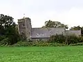 Low building with tiled roof and non-vertical square tower, surrounded by trees and with grass in the foreground.