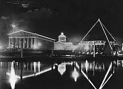 Greek architecture pictured at night in black in white. The foreground is occupied by a small body of water and the building in the middle and background have their perimeter outlined with string lights.