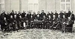 A photograph of a group of men in a semi-circle consisting of two rows. The front row is seated. There is a desk in the middle of the semi-circle with a man seated behind it.