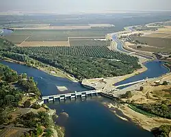 View from the air of a river flowing through the center from right to left; a dam blocks the water flow in the right half presumably to send some of the water to a canal that leads off to the upper left.